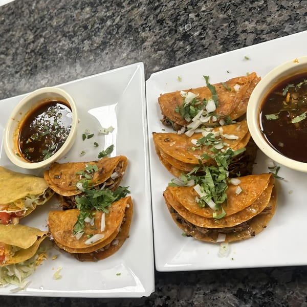 Two plates of birria tacos with cilantro, onions, and dipping sauce on a dark countertop.