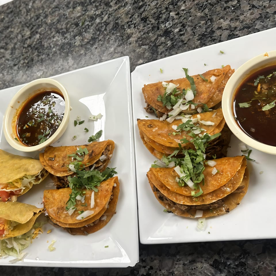Two plates of birria tacos with cilantro, onions, and dipping sauce on a dark countertop.